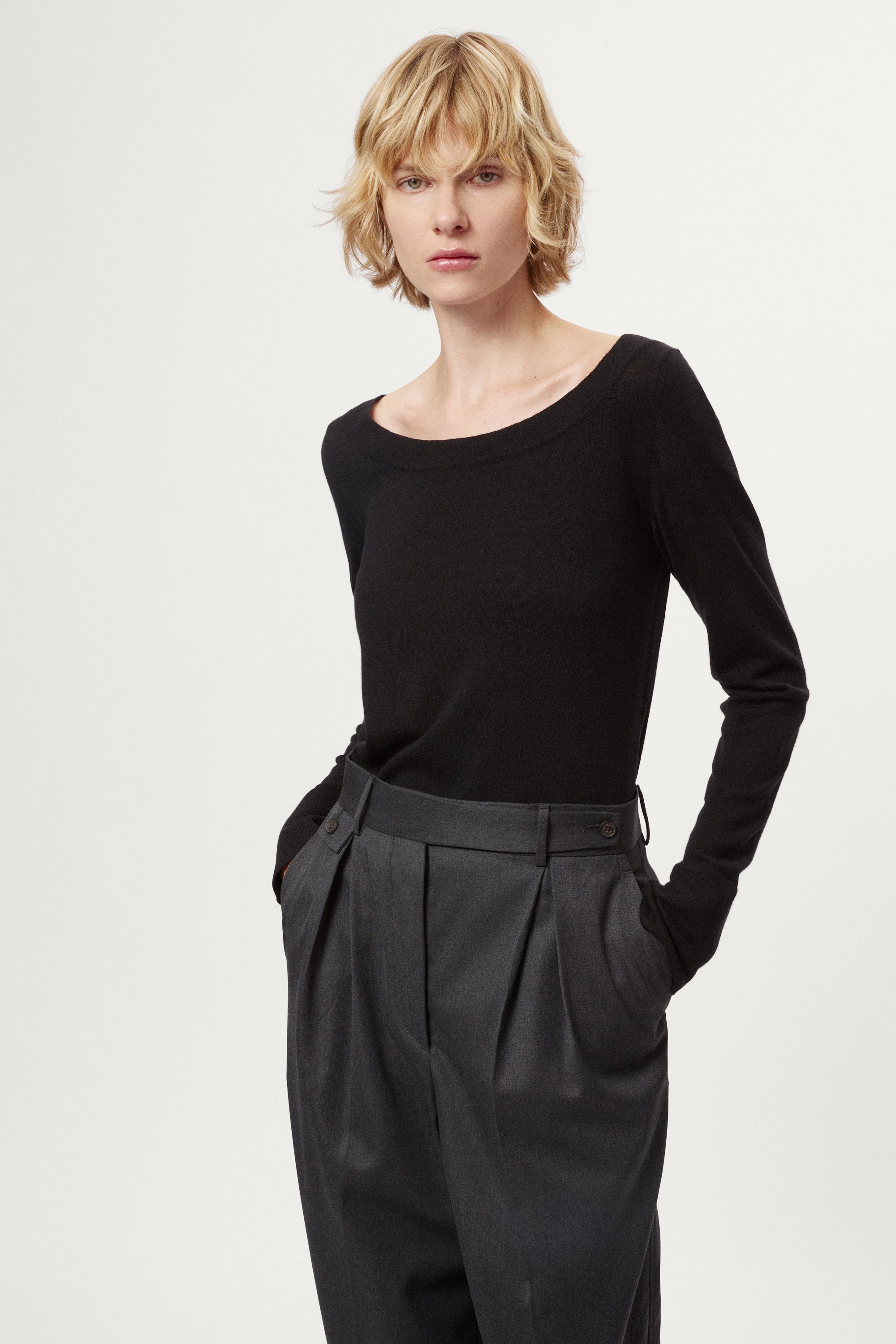 Person wearing a black long-sleeve top and dark gray pleated pants on a white background