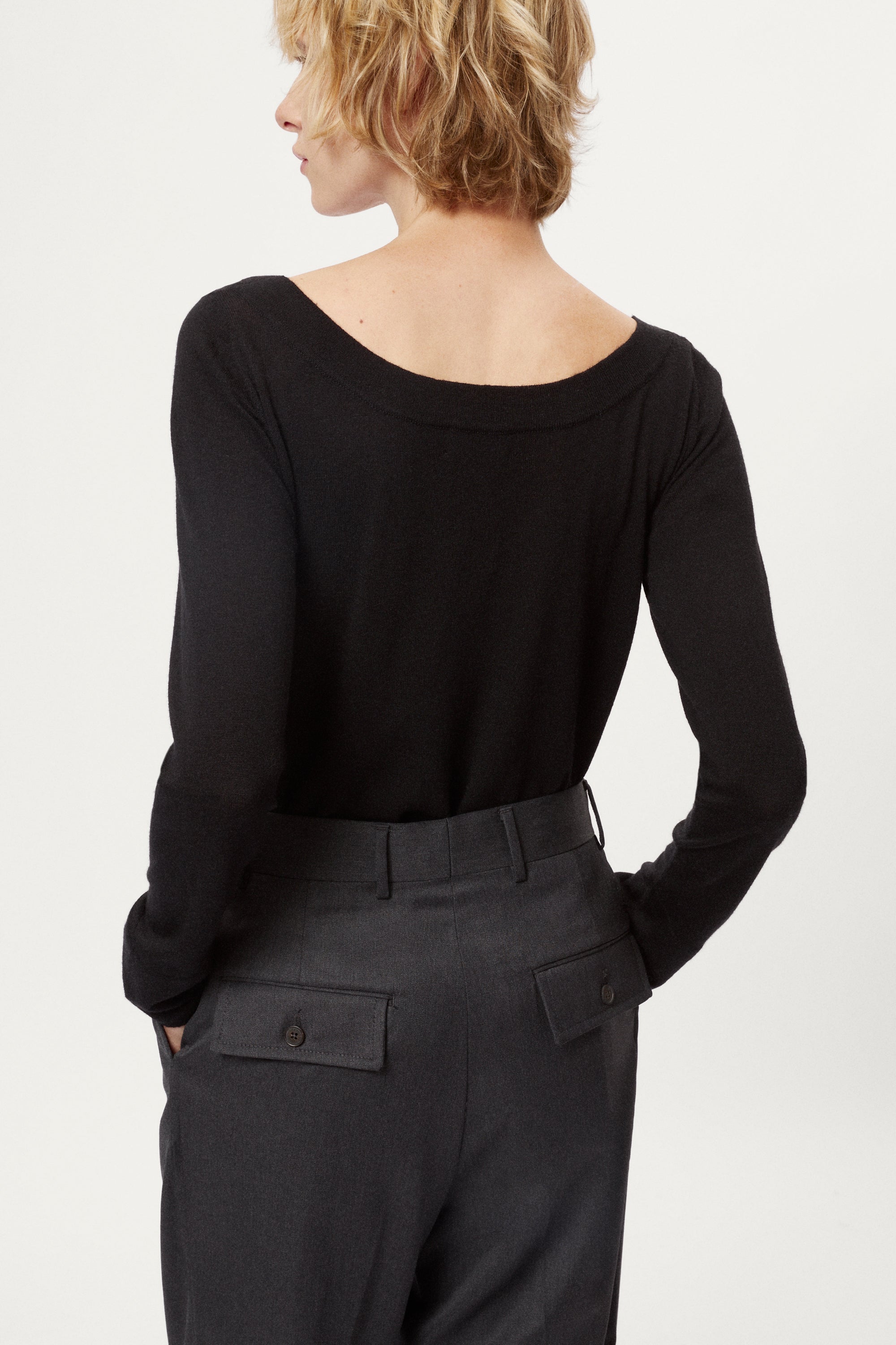 Person wearing a black long-sleeve top and dark gray pleated pants on a white background