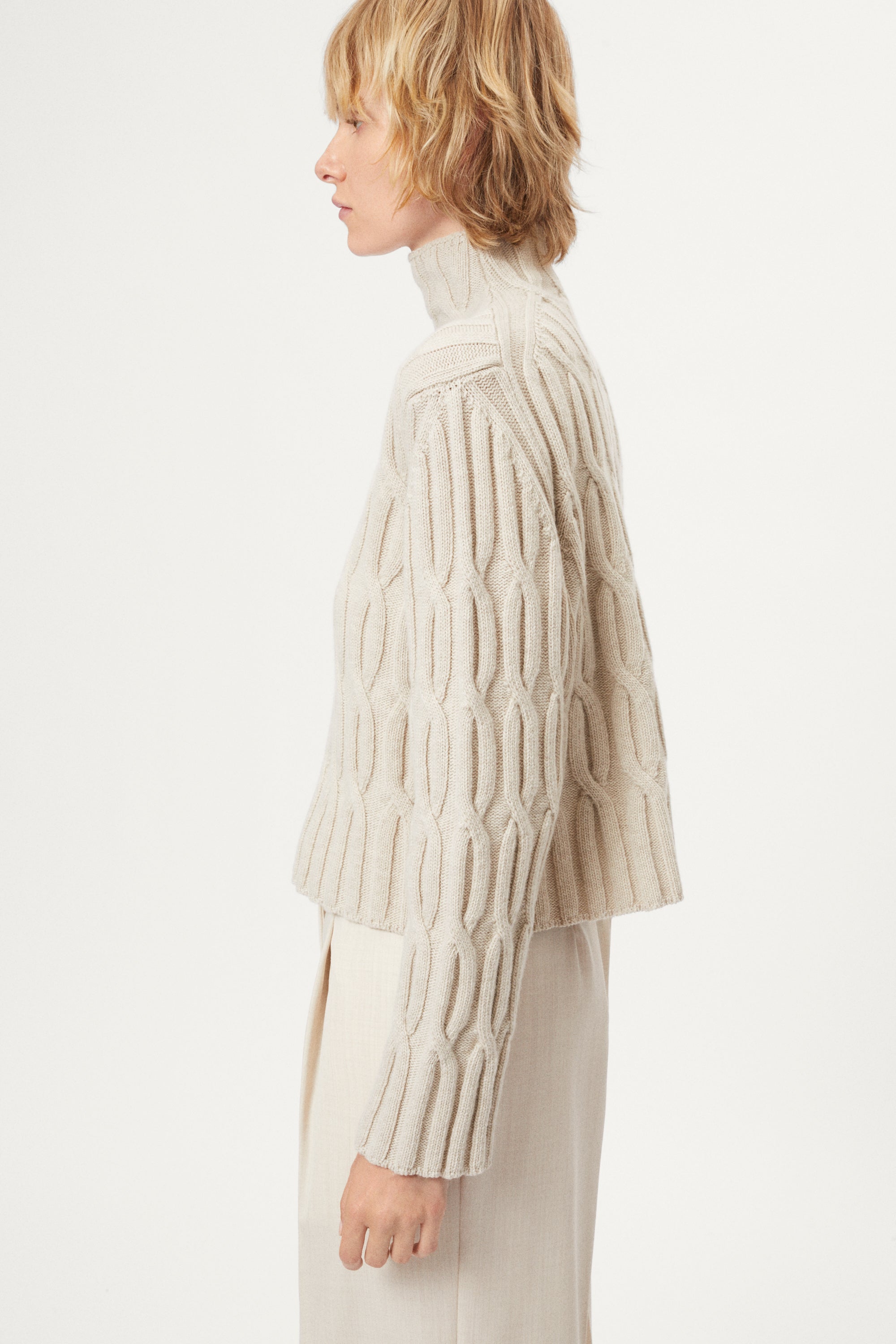 woman wearing a woolen  beige cable knit turtleneck sweater on a white background