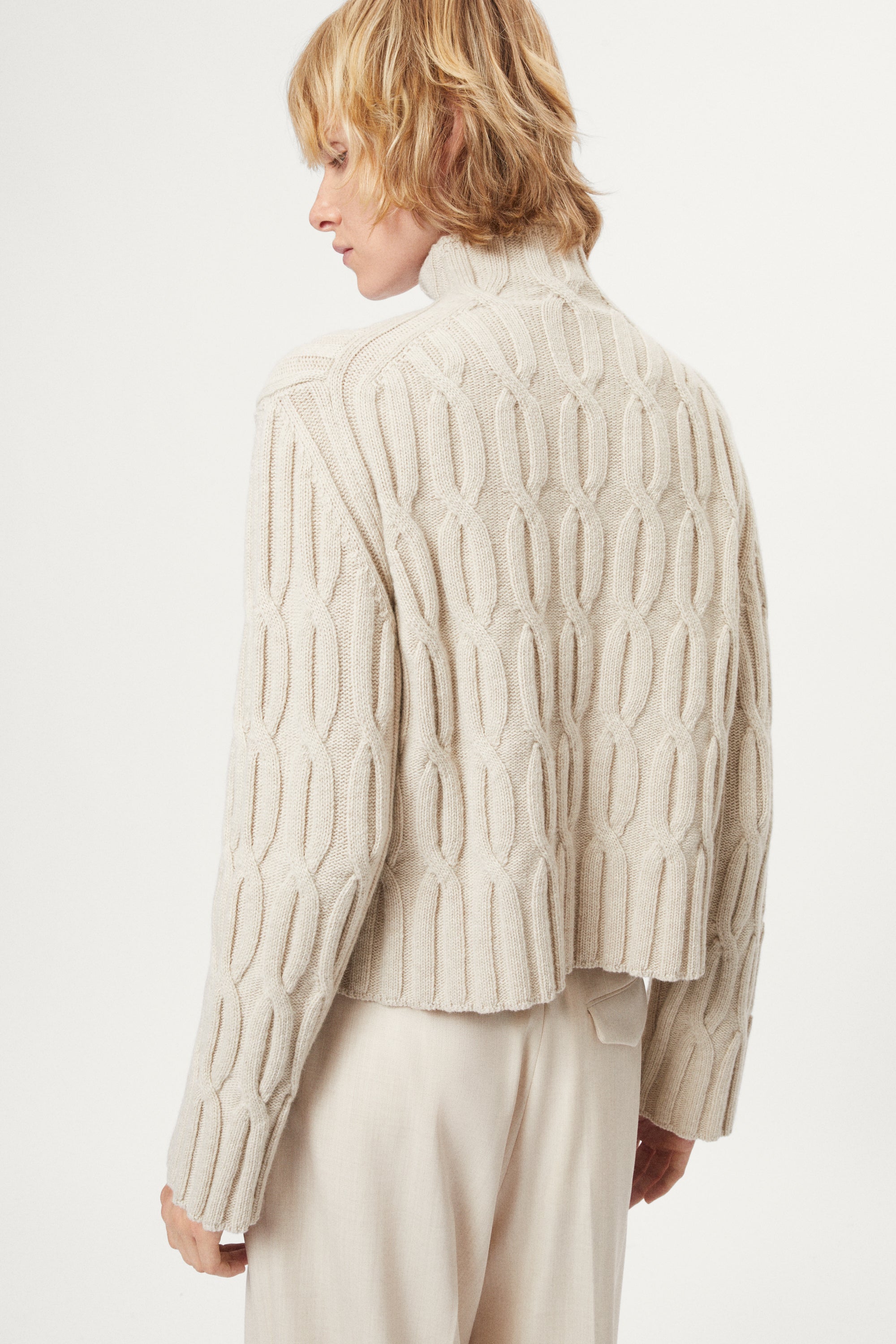 woman wearing a woolen  beige cable knit turtleneck sweater on a white background