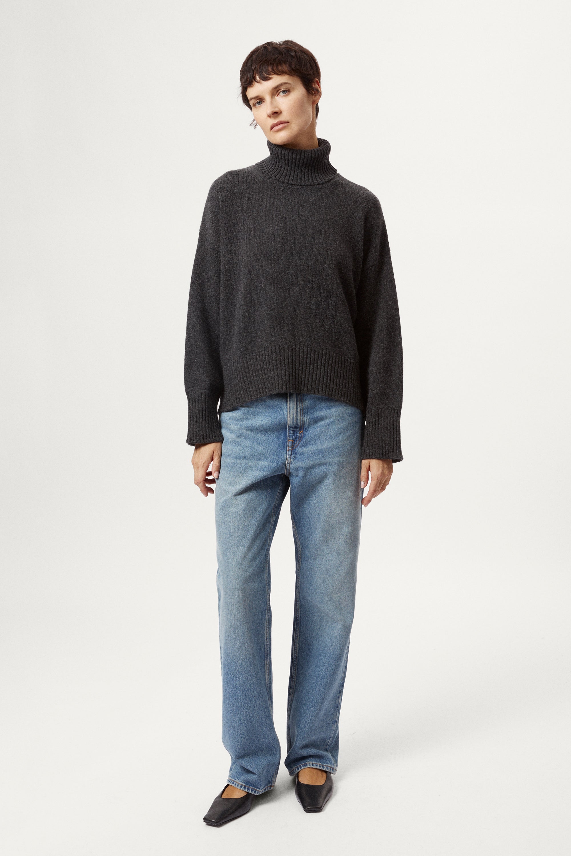 woman wearing a woolen dark grey turtleneck sweater and blue jeans on a white background