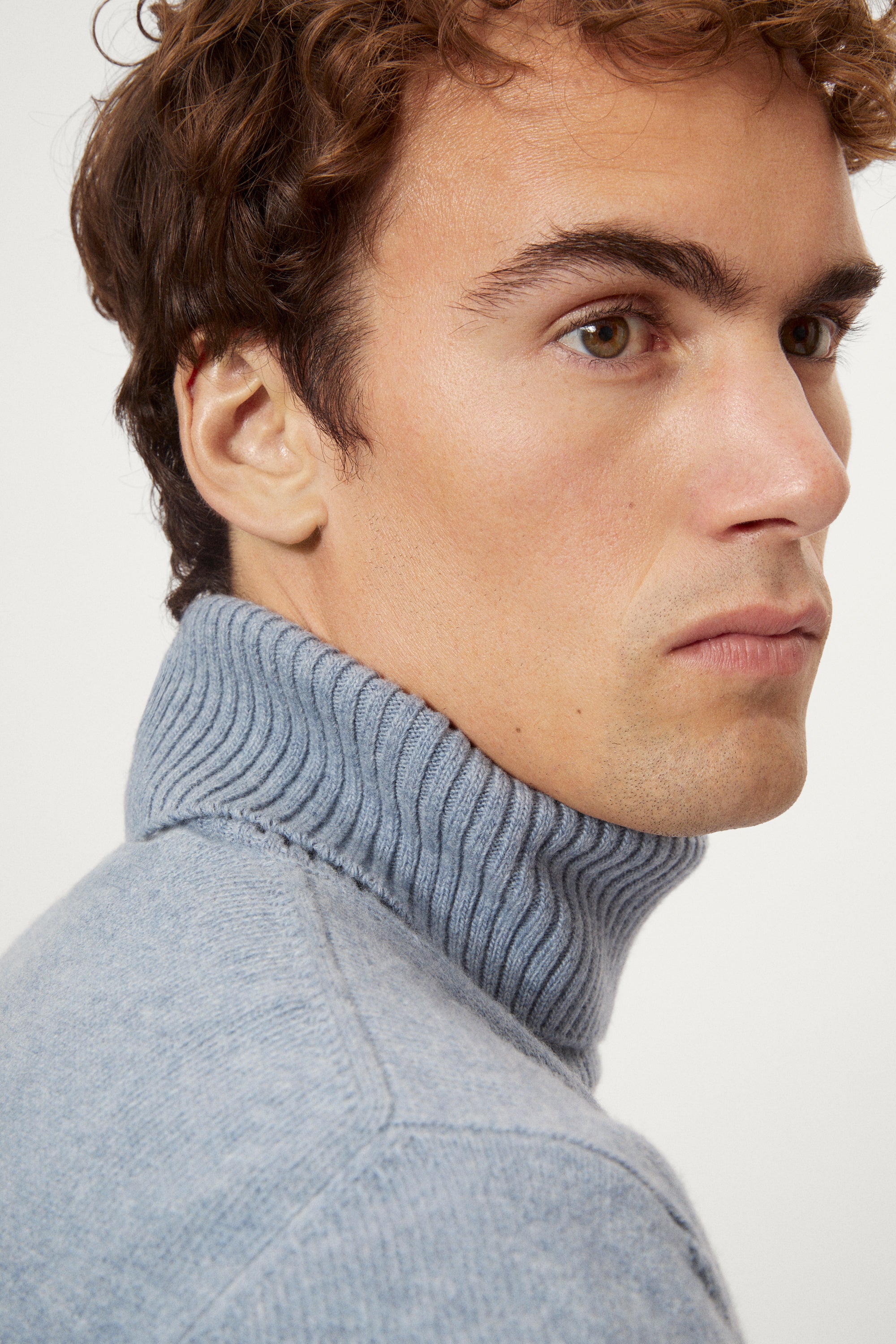 Man wearing a woolen light blue turtleneck sweater and brown pants on a white background