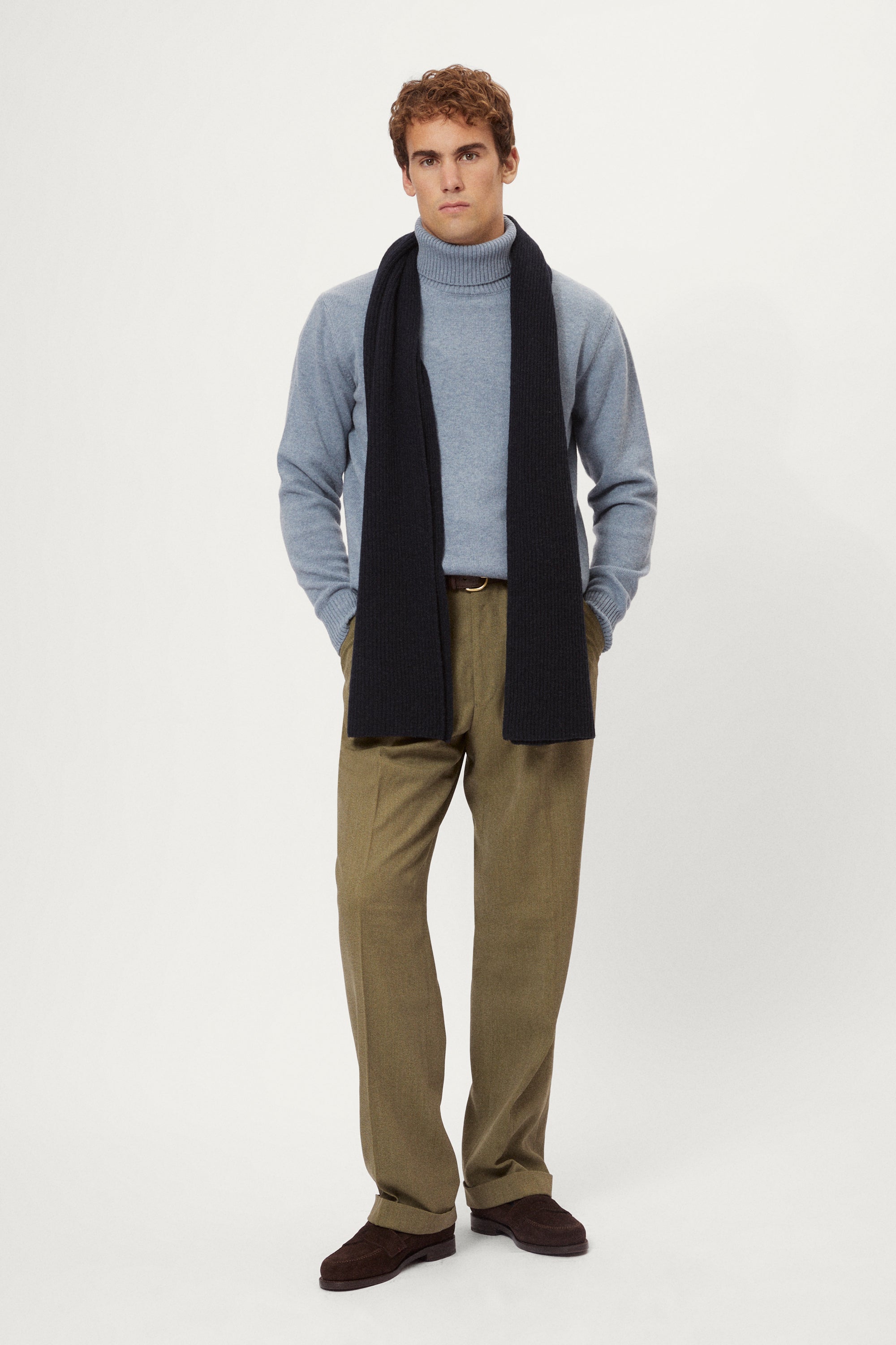 Man wearing a woolen light blue turtleneck sweater and brown pants on a white background