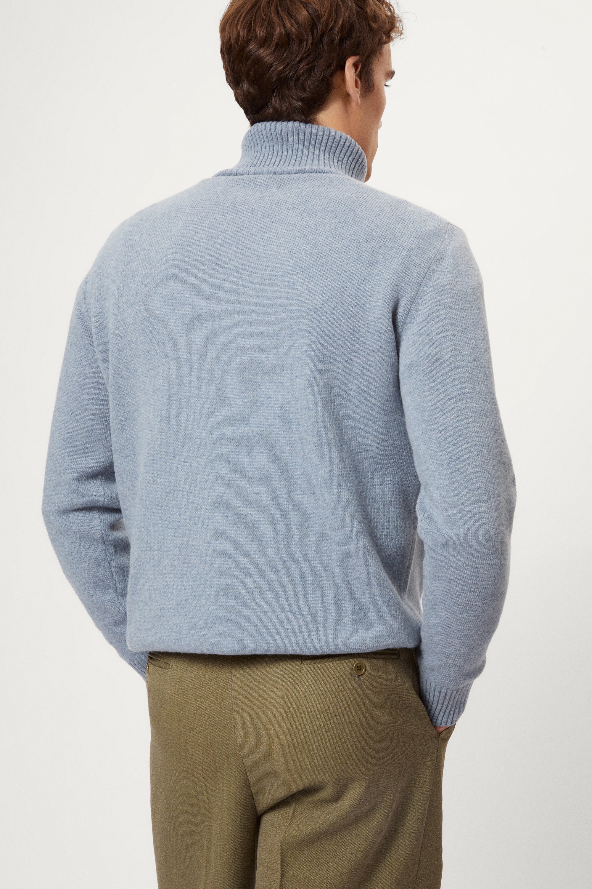 Man wearing a woolen light blue turtleneck sweater and brown pants on a white background