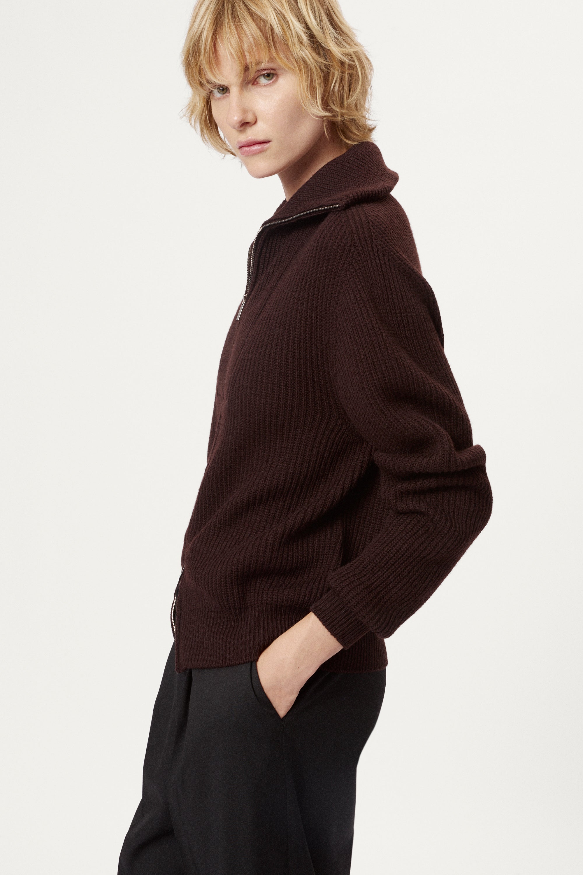 woman wearing a dark brown zip-up sweater and grey pants on a white background