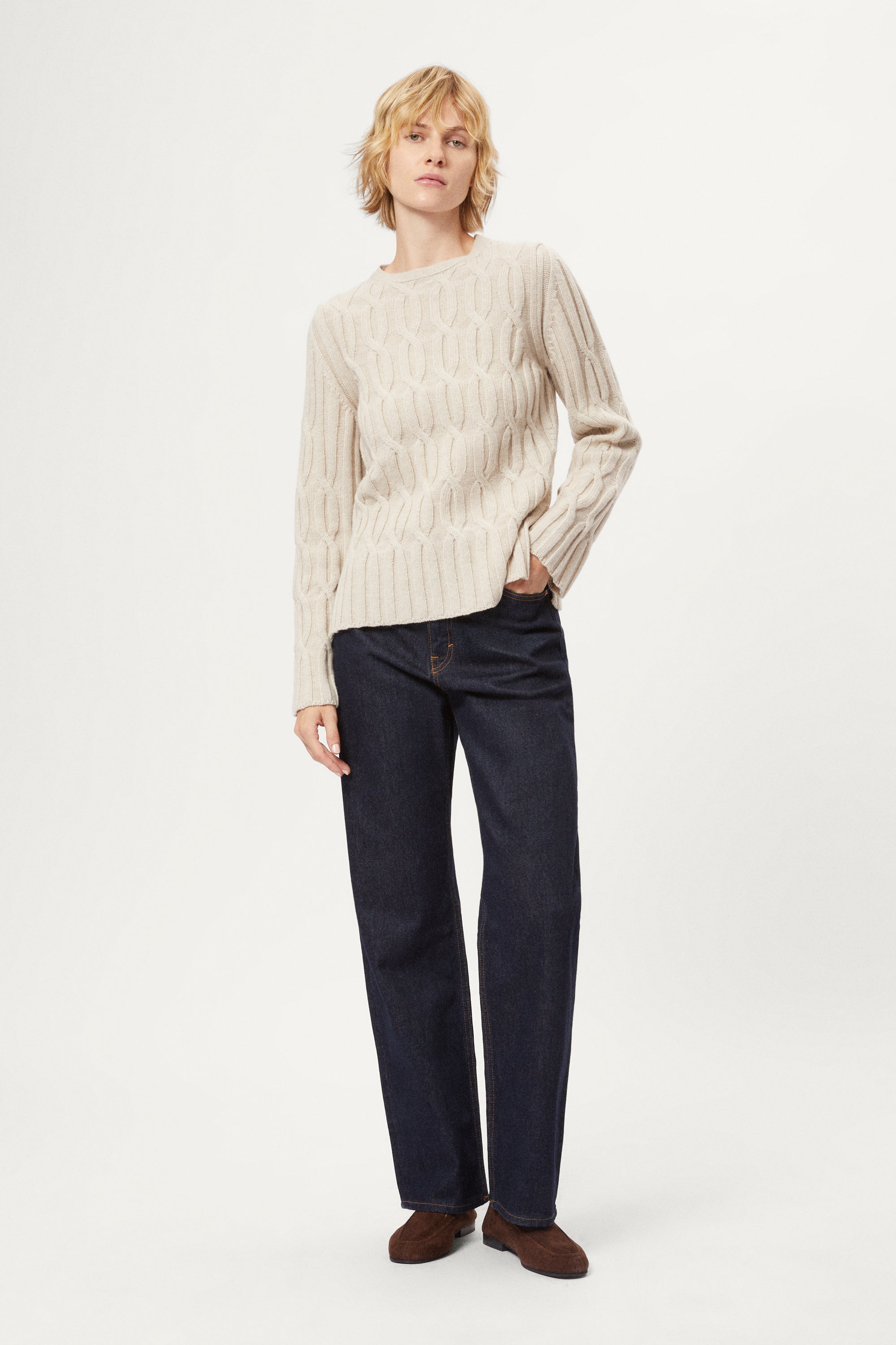 Woman wearing an ecru woolen cable knit sweater and dark jeans on a white background