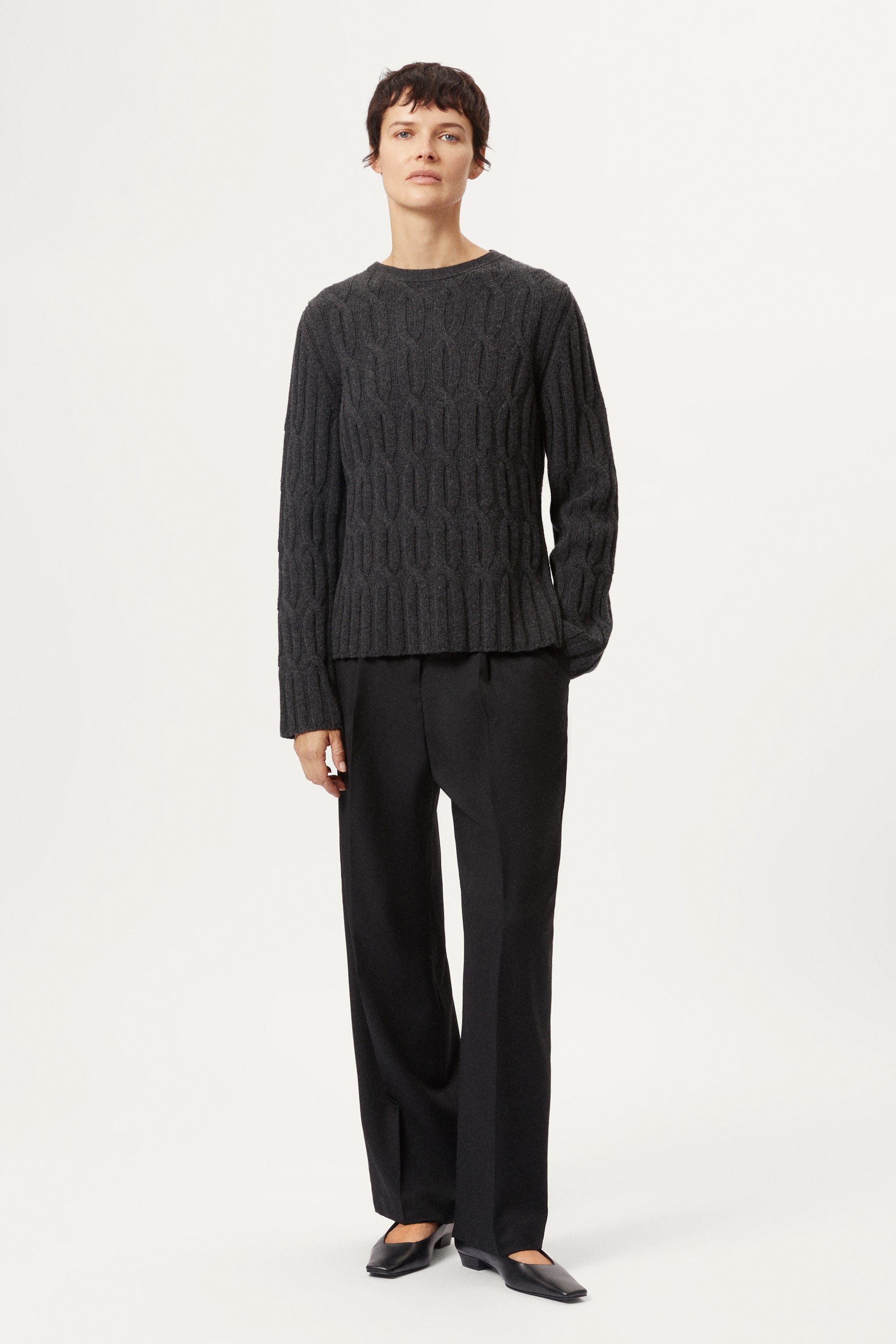 Woman wearing a woolen dark gray cable knit sweater and grey pants on a white background