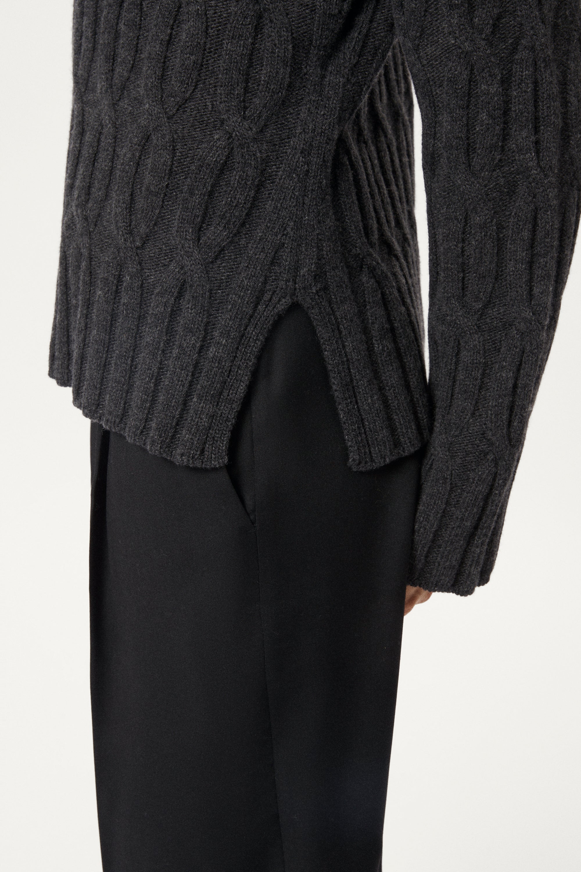 Woman wearing a woolen dark gray cable knit sweater and grey pants on a white background