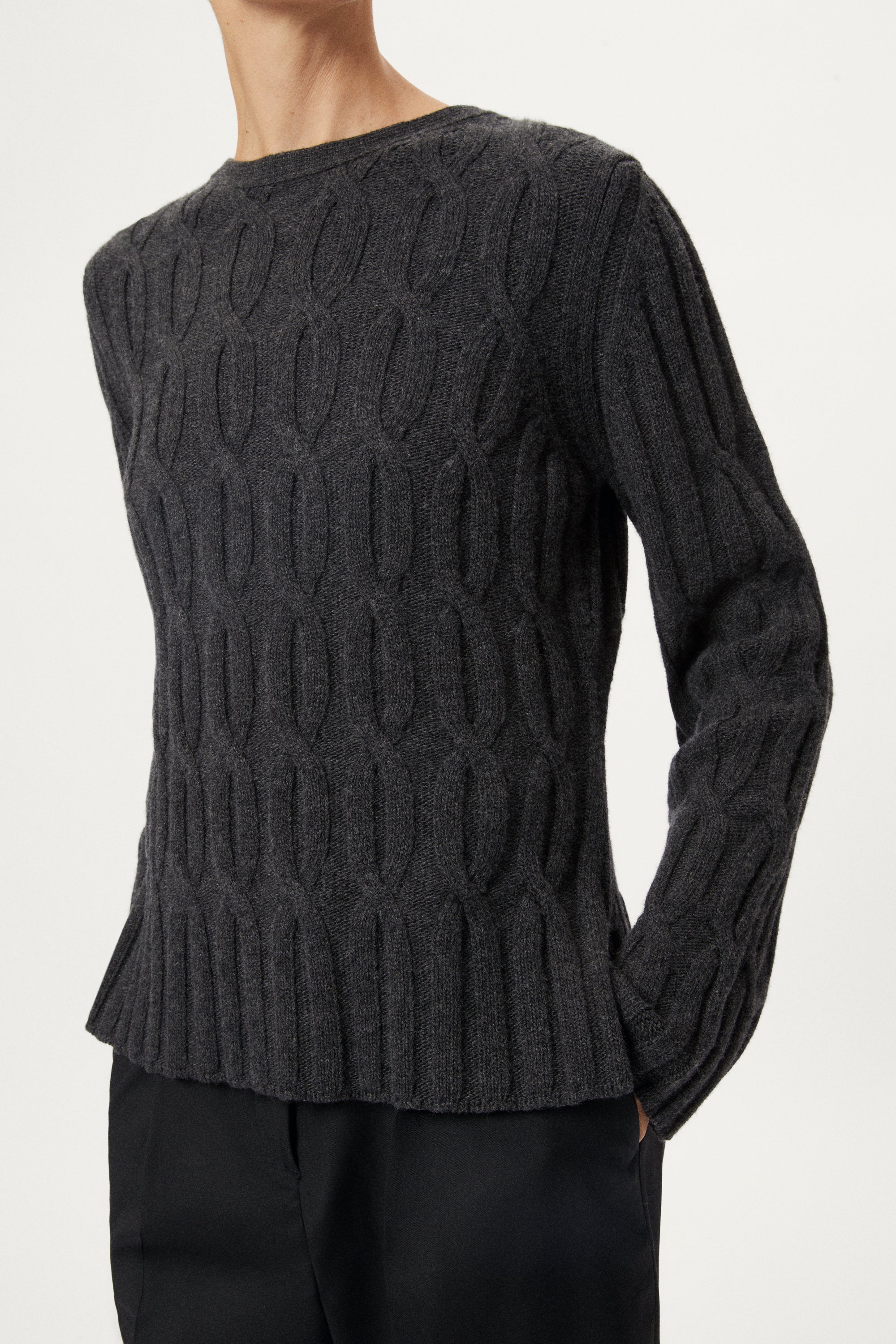 Woman wearing a woolen dark gray cable knit sweater and grey pants on a white background