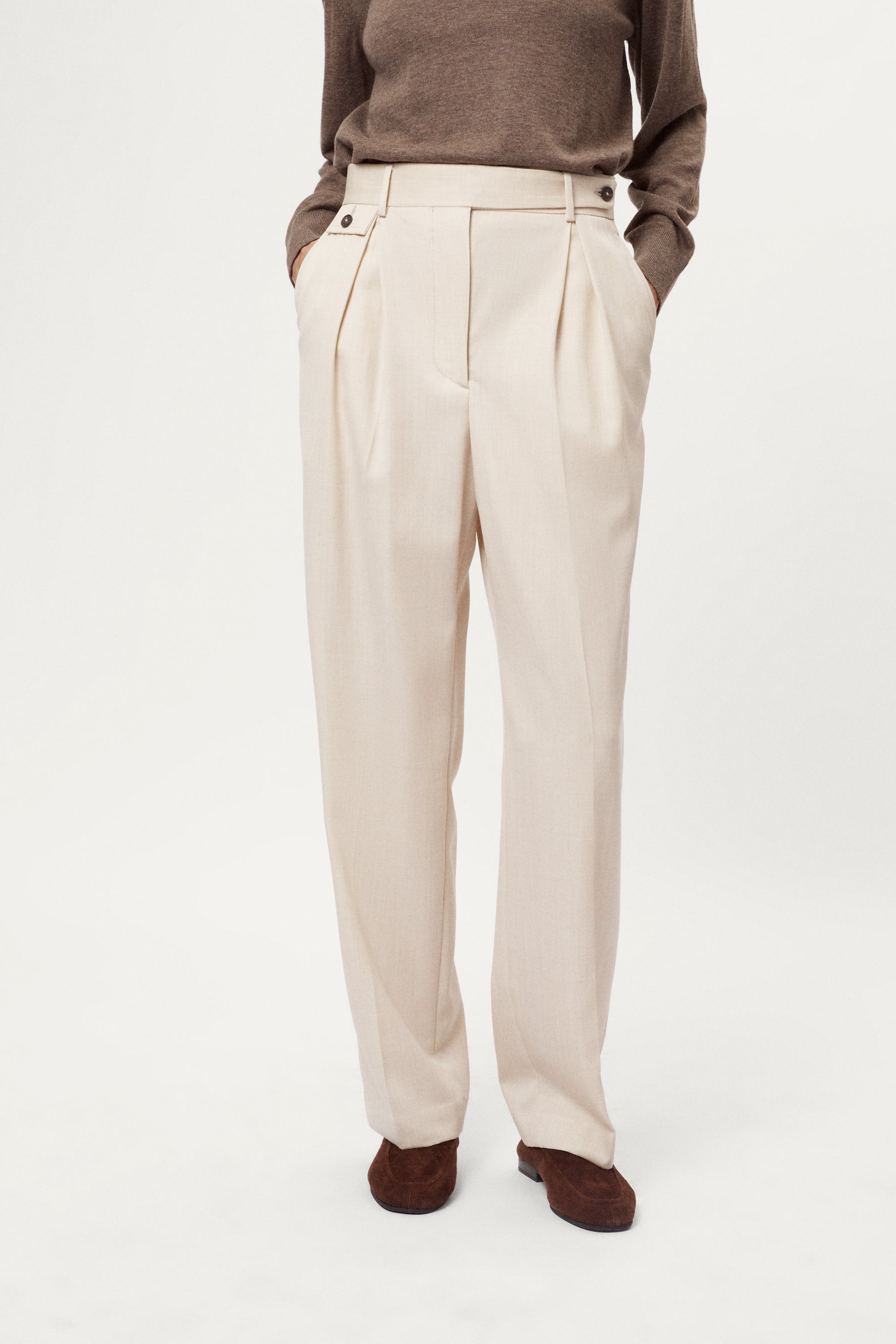 Beige pleated pants worn with a brown sweater on a white background