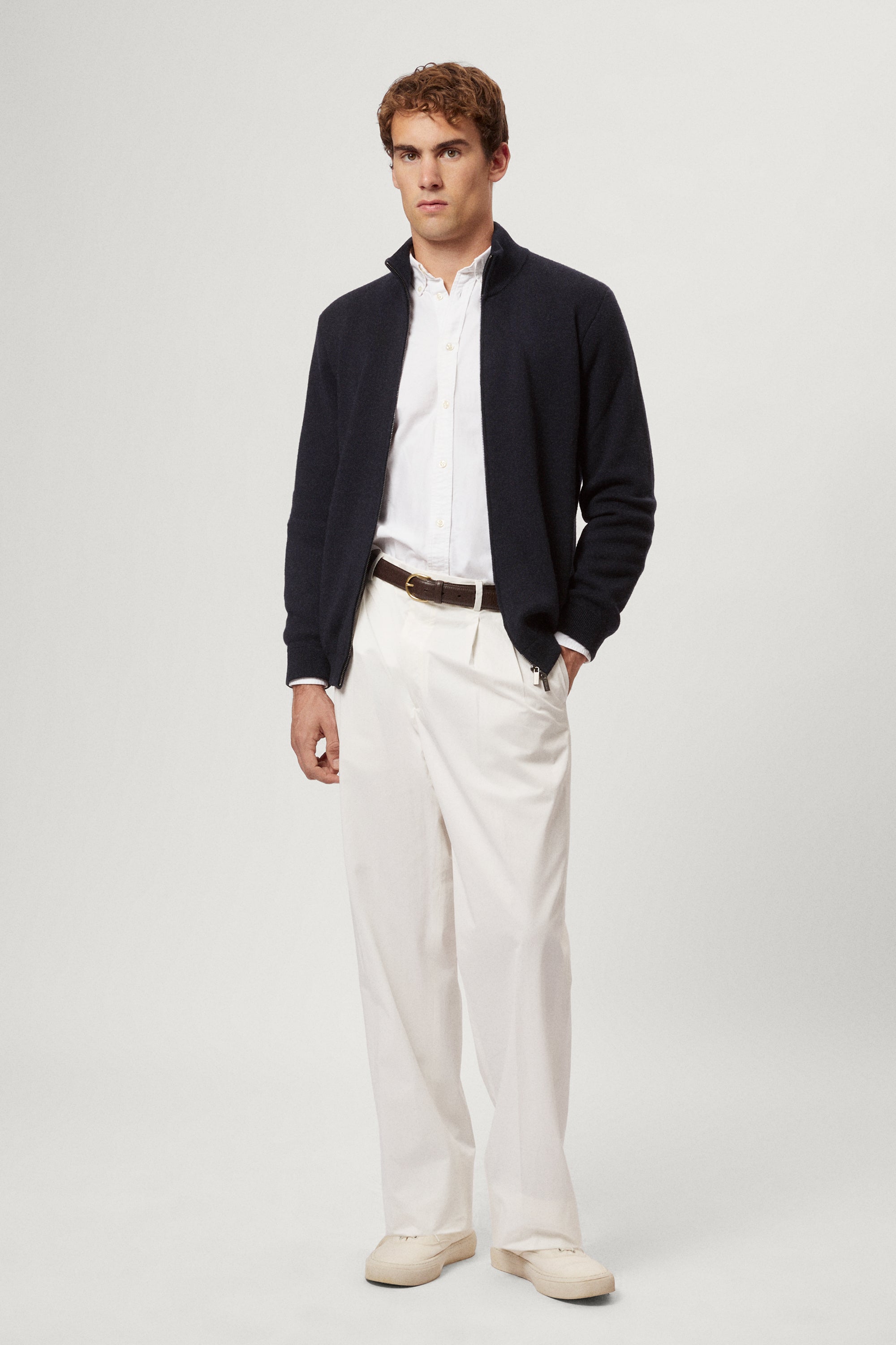 Man wearing a navy blue zip-up sweater over a white shirt with light-colored pants on a plain background