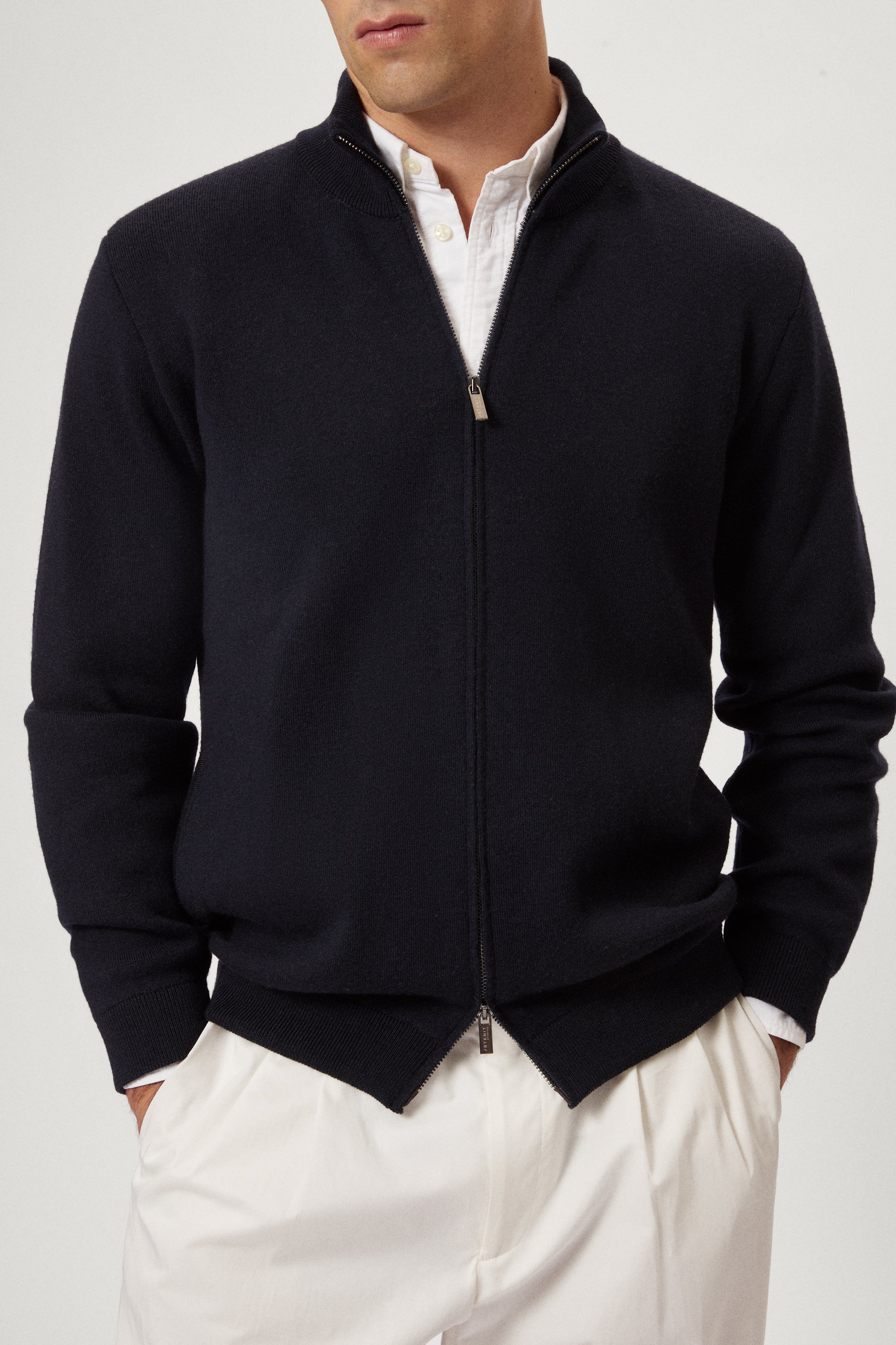 Man wearing a navy blue zip-up sweater over a white shirt with light-colored pants on a plain background