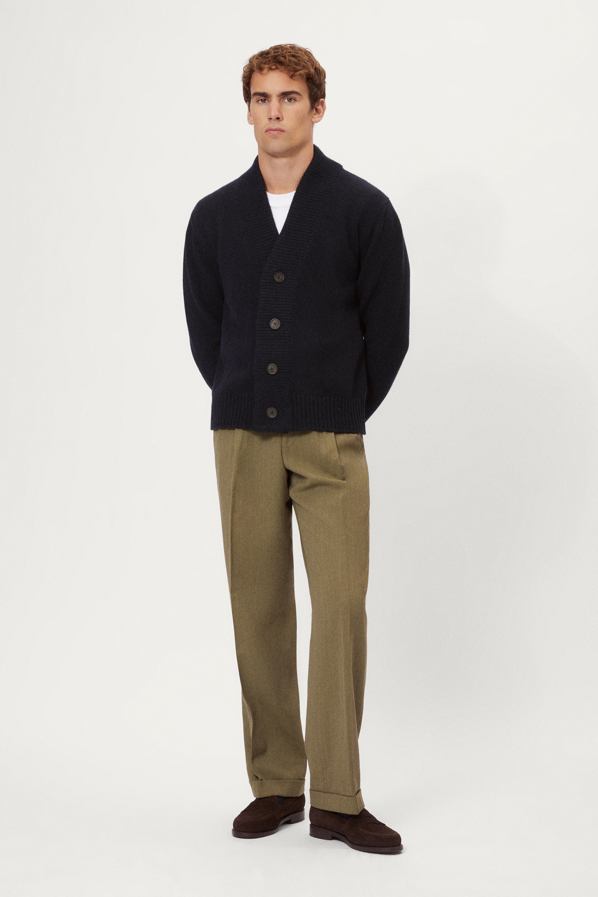 Man wearing a woolen blue cardigan over a white shirt with beige pants on a white background