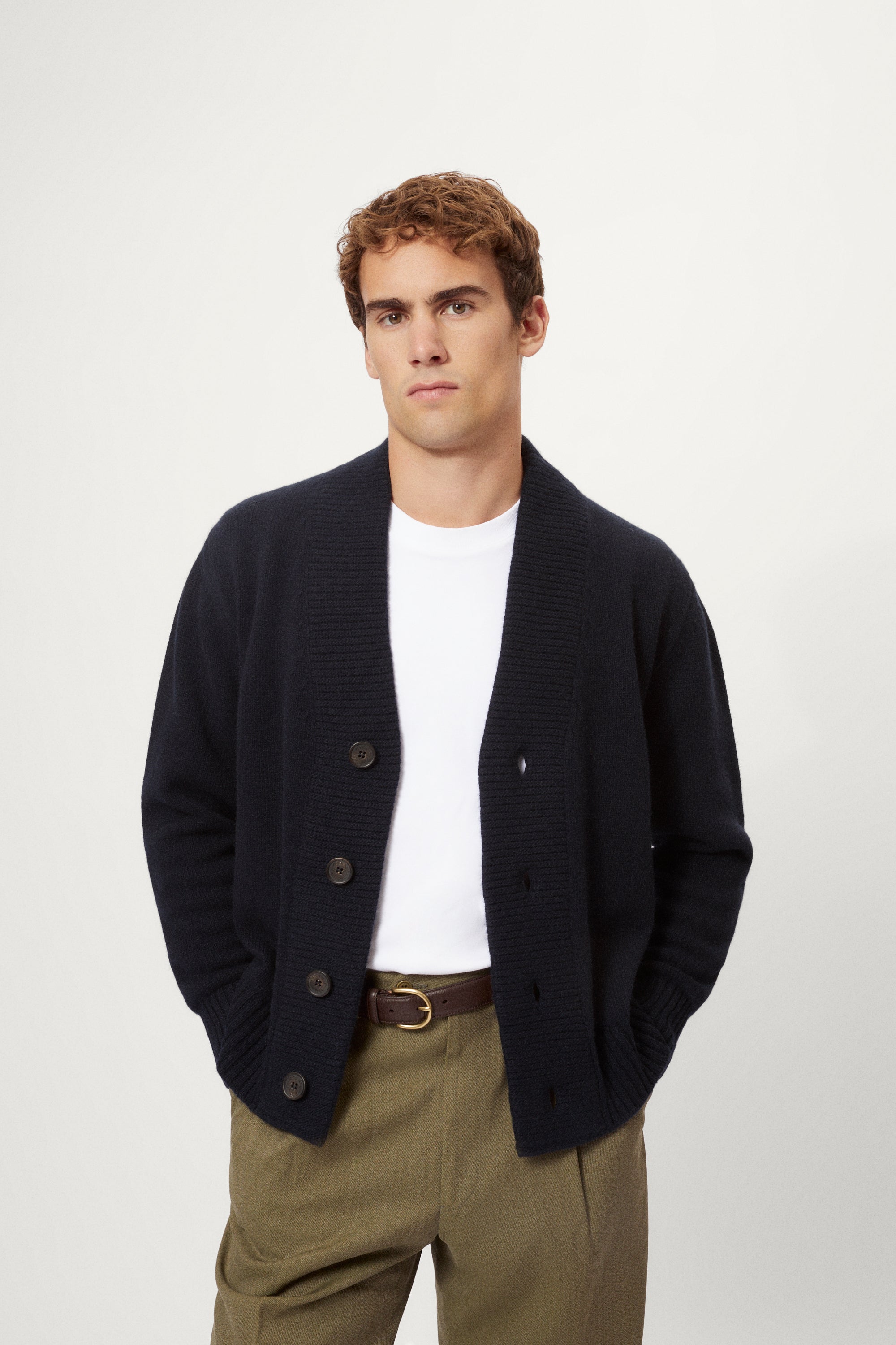 Man wearing a woolen blue cardigan over a white shirt with beige pants on a white background