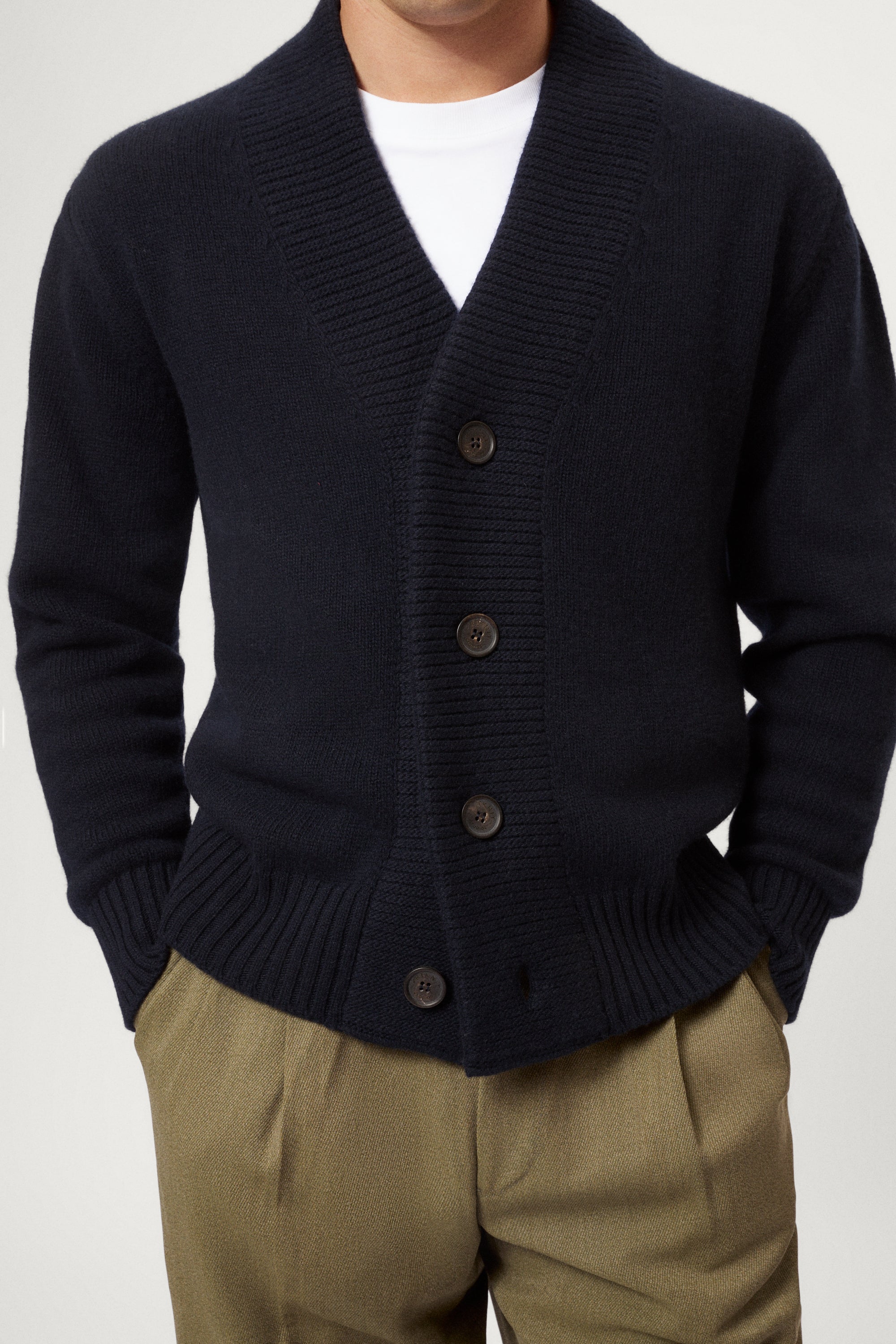 Man wearing a woolen blue cardigan over a white shirt with beige pants on a white background