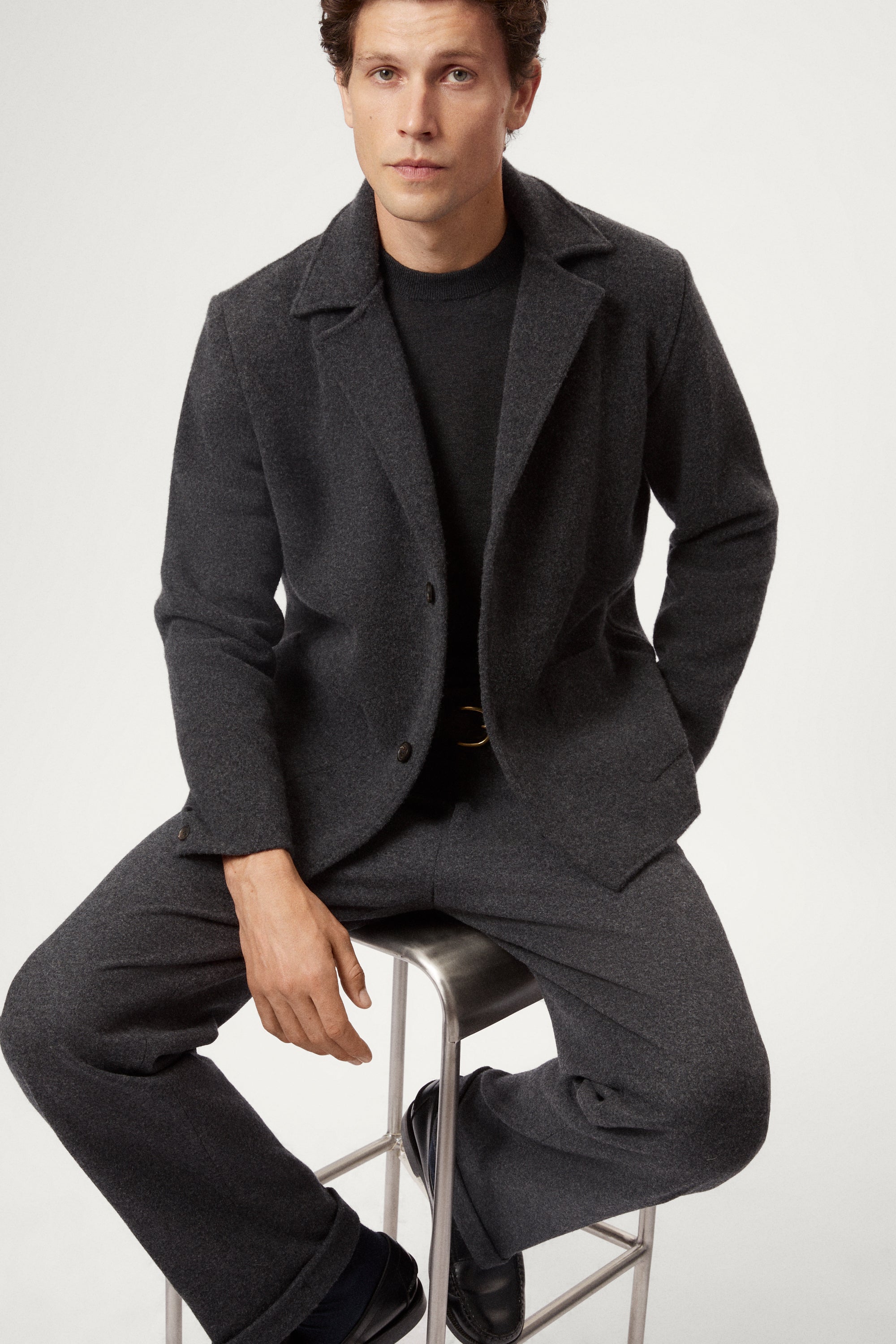 Man wearing a Woolen dark gray Jacket and pants sitting on a stool against a white background