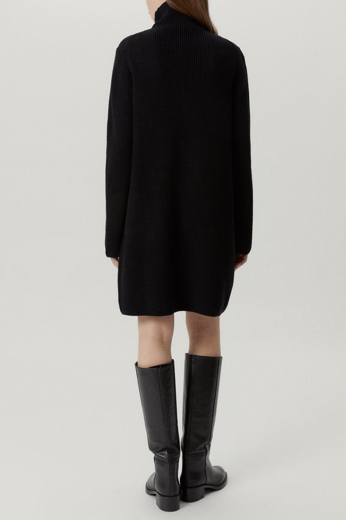 Person wearing a black turtleneck dress and knee-high boots on a light gray background