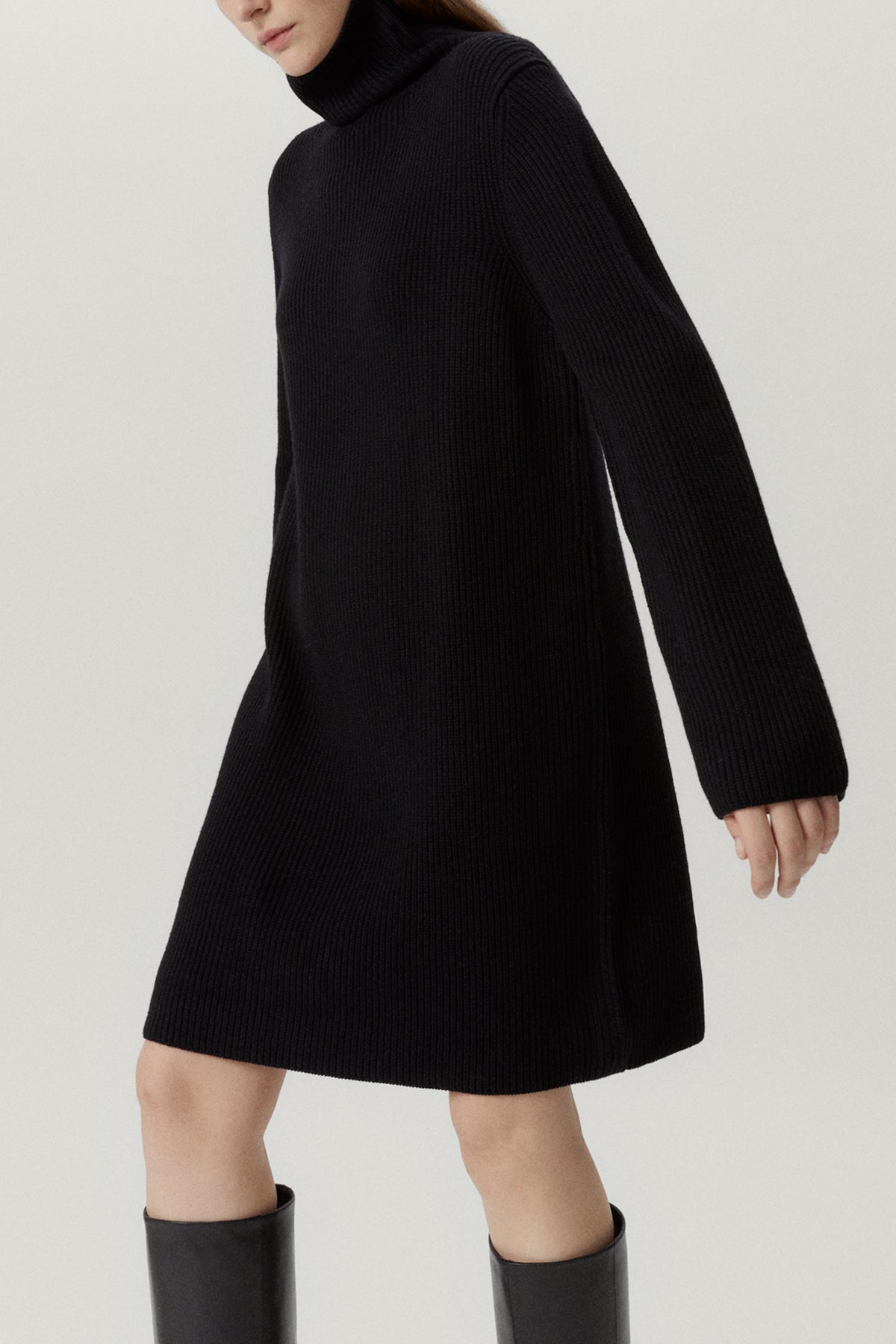 Person wearing a black turtleneck dress and knee-high boots on a light gray background